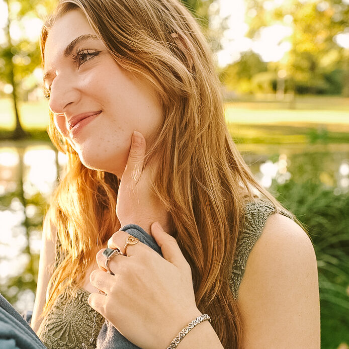 A young woman with long, wavy hair lightly touches her cheek while smiling, set against a blurred outdoor background with greenery and a pond.