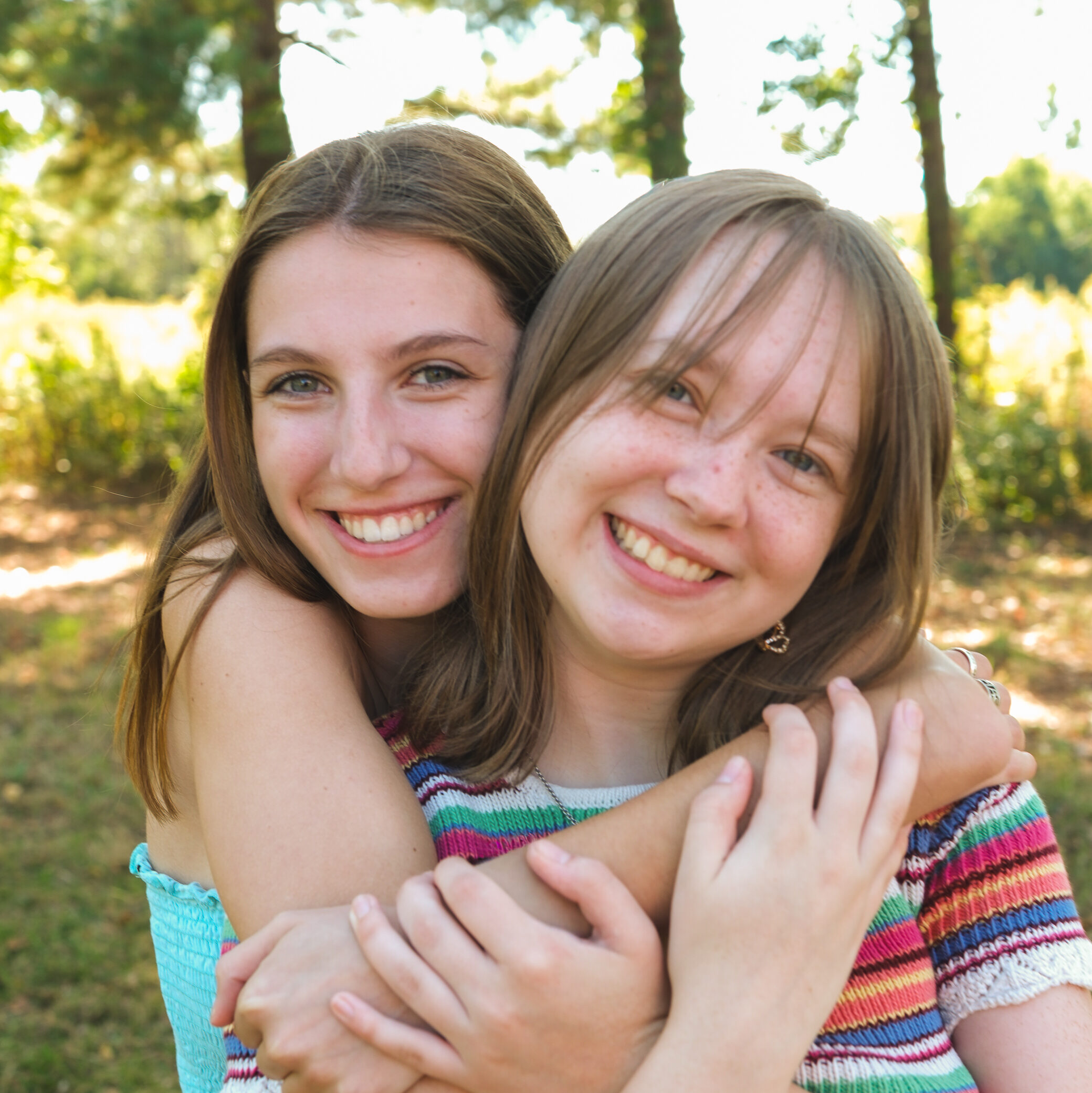 Two young women embracing each other, both smiling warmly, standing outdoors in a natural setting with trees in the background.