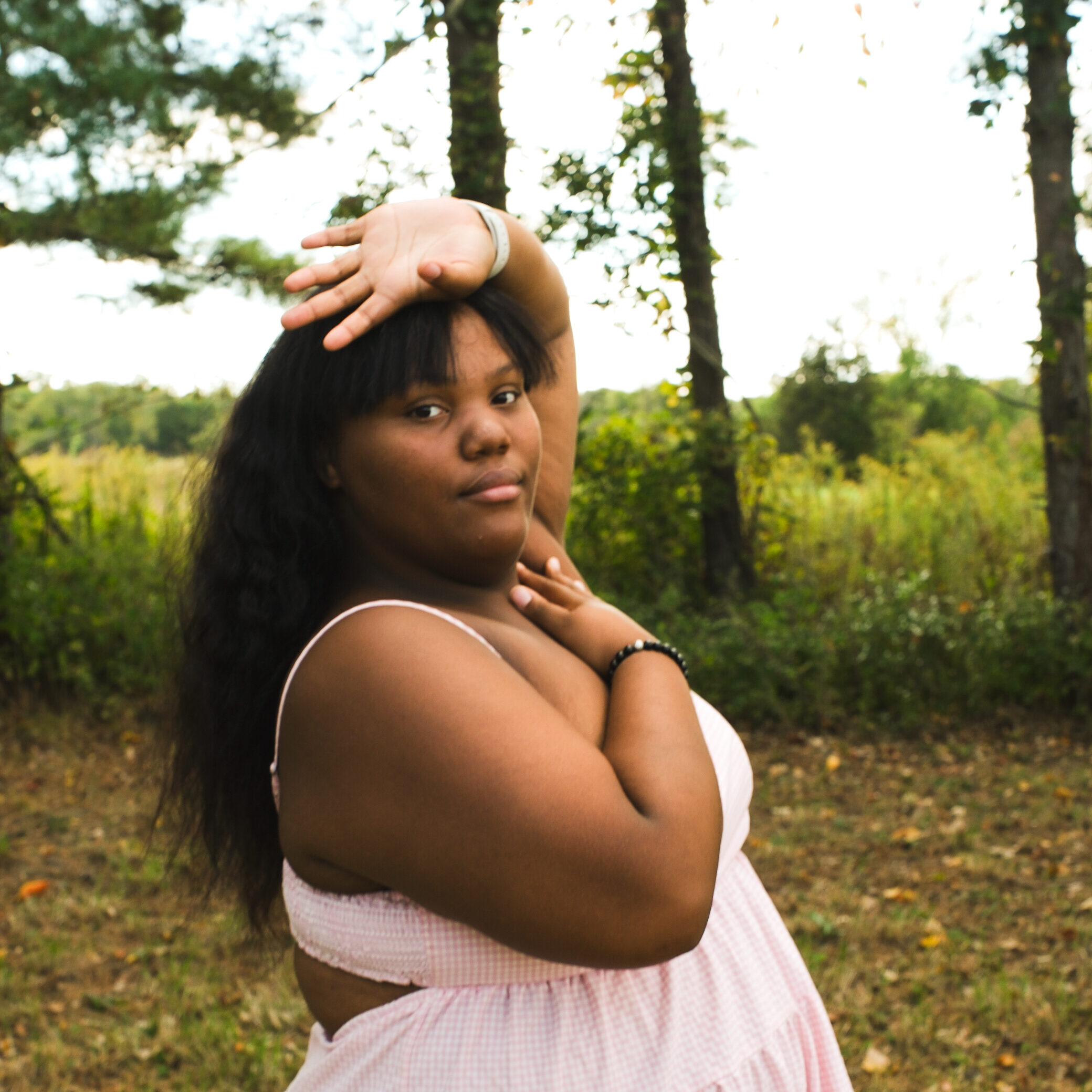 A young woman stands with her arms framing her face in a pink dress