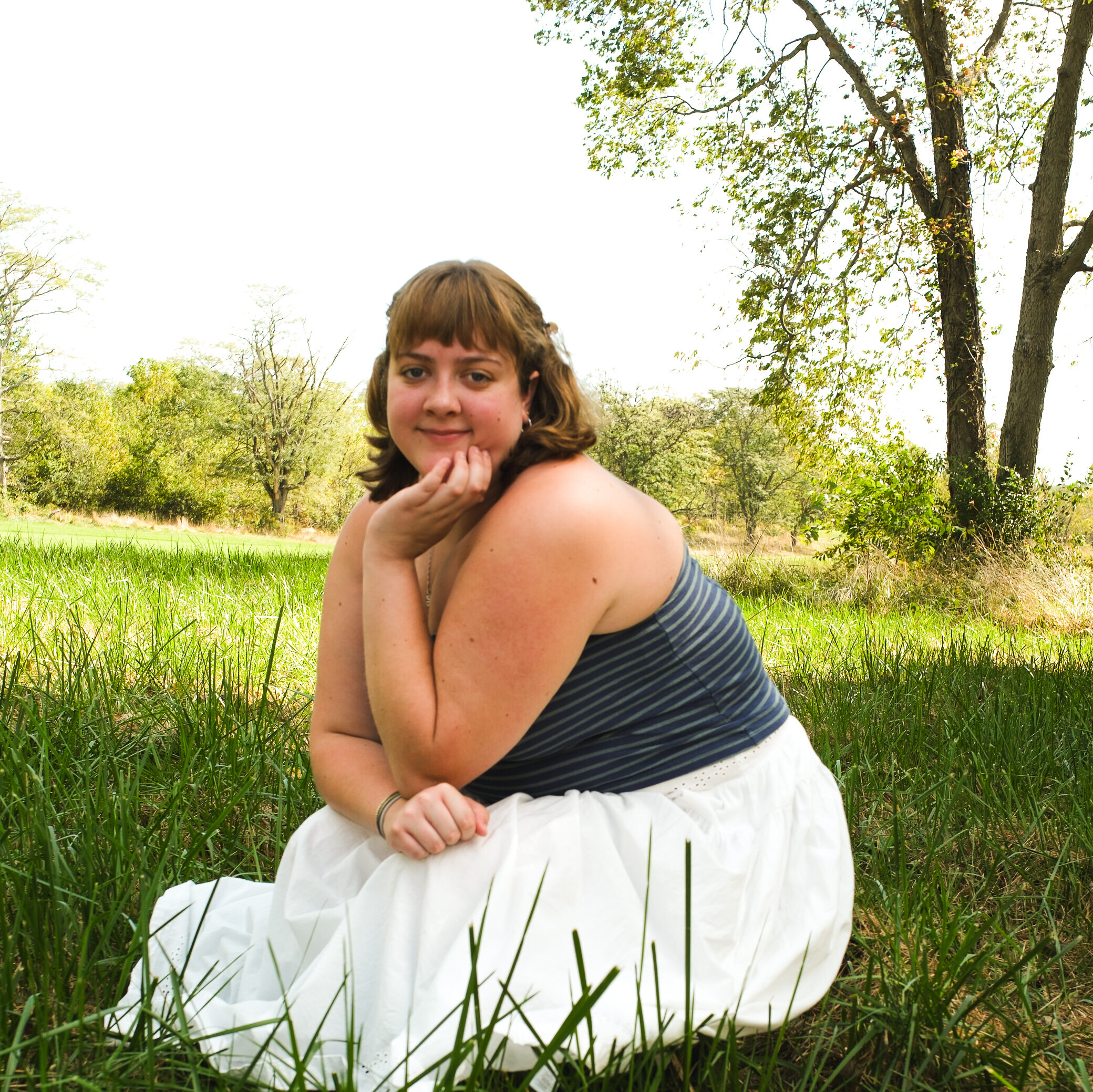 a young woman is crouched on the ground with her head leaning gently on her hand, looking at the camera

