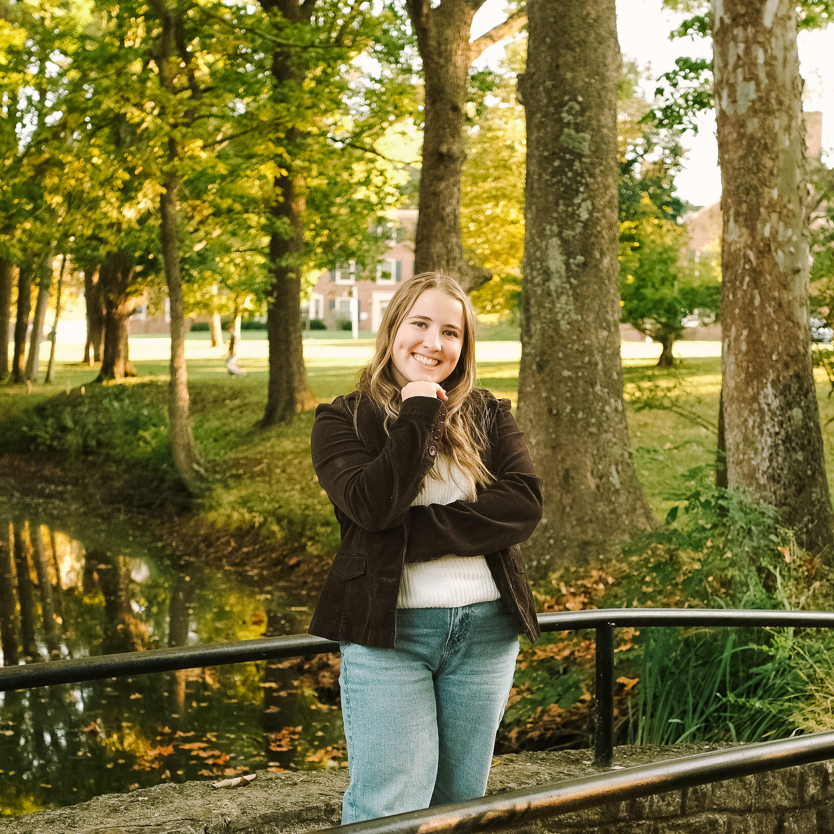 A young woman stands smiling on a bridge surrounded by lush green trees and a calm waterway, wearing a brown jacket and light blue jeans.