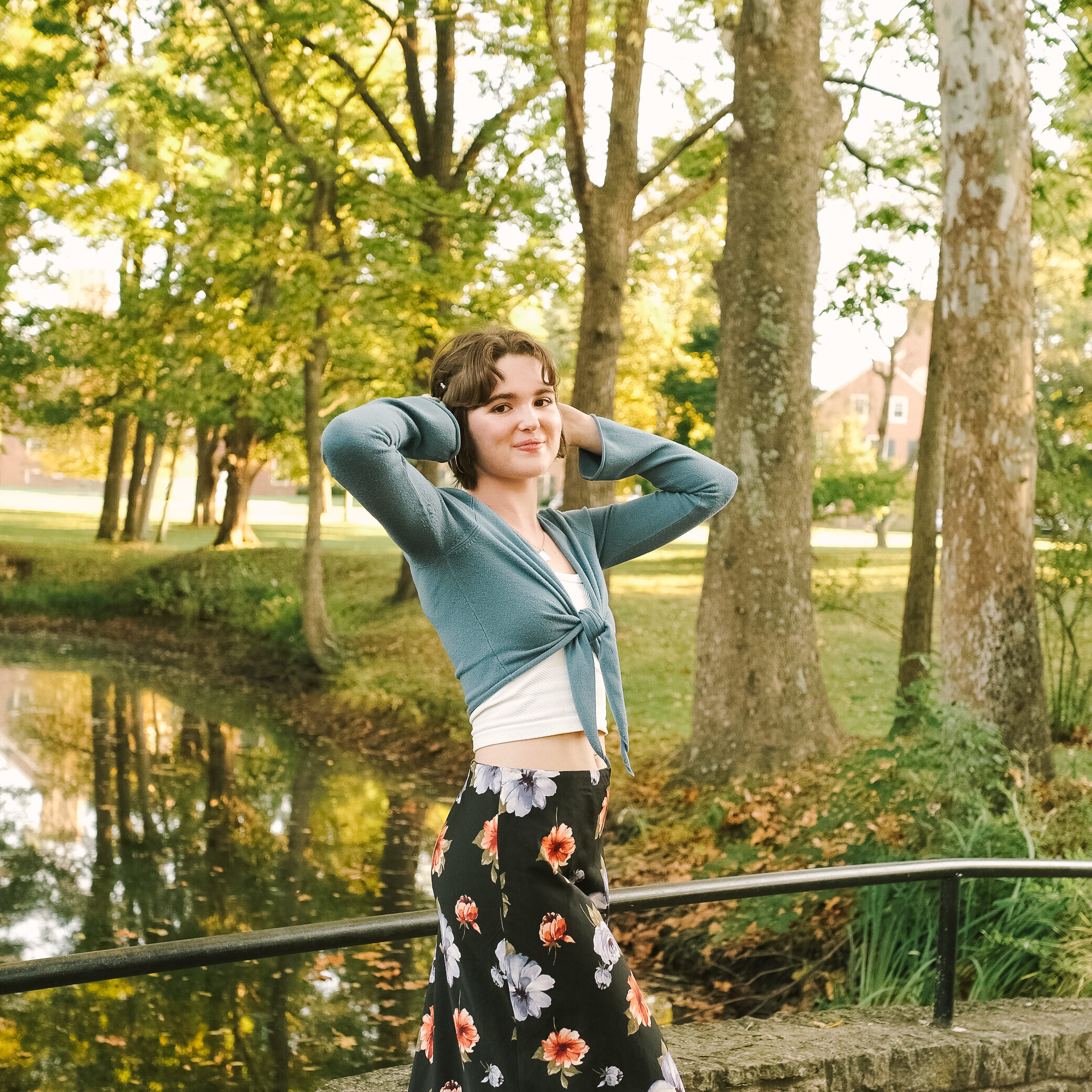 A young woman stands on a bridge by a serene pond, surrounded by trees. She has shoulder-length hair, wears a blue cardigan, and a floral skirt, with her hands behind her head, smiling at the camera.